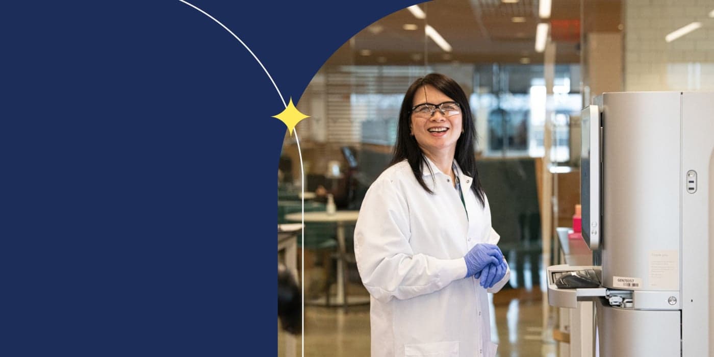 A person in a white lab coat, safety goggles, and blue latex gloves standing in a lab smiling at the camera. They have shoulder-length black hair, and they are standing in front of a face-height screen and keyboard.